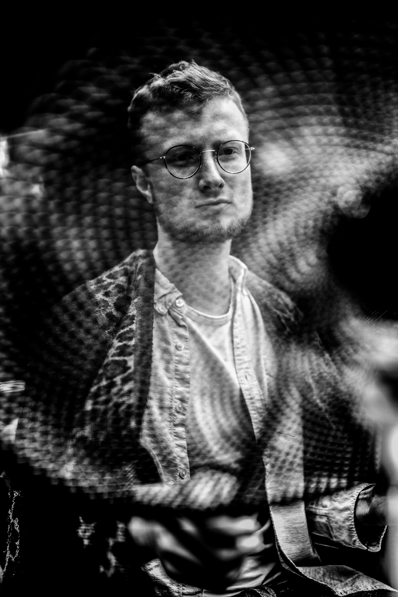Mann mit Brille und Hemd ist in einer Schwarzweißaufnahme durch ein gemustertes, unscharfes Vordergrundobjekt zu sehen.