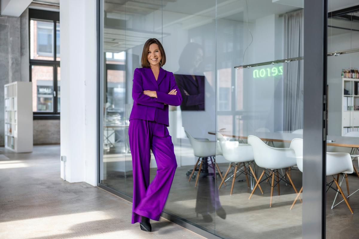 Portrait von Vivian Stürmann in ihrer Agentur Zeron. Sie trägt einen lila Hosenanzug und lehnt an einer Glasfront.
