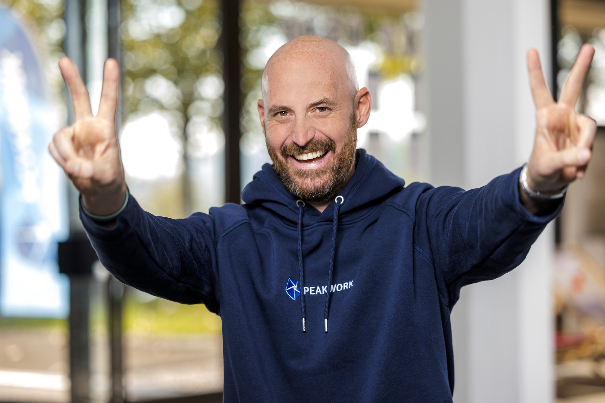 Mann in blauem Hoodie zeigt drinnen beide Hände mit Peace-Zeichen und lächelt.