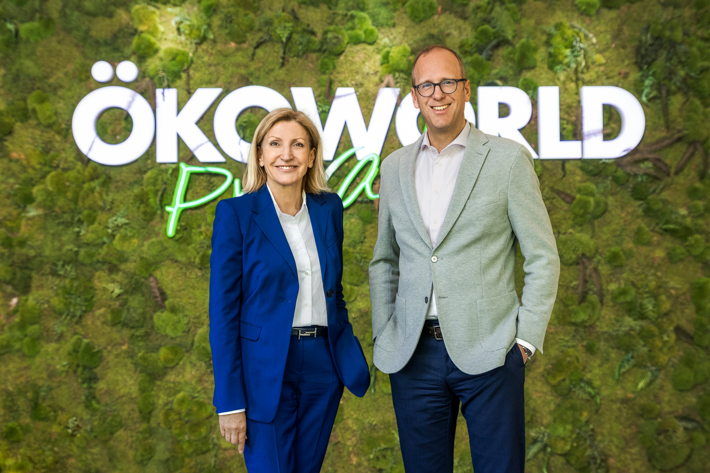 Geschäftsführung von Ökoworld. Eine Dame steht im blauen Hosenanzug mit einer Hand in der Hosentasche. Der Mann trägt ein graues Jacket und ein weißes Hemd. Beide stehen vor einer grünen Moos Wand und schauen lächelnd in die Kamera.