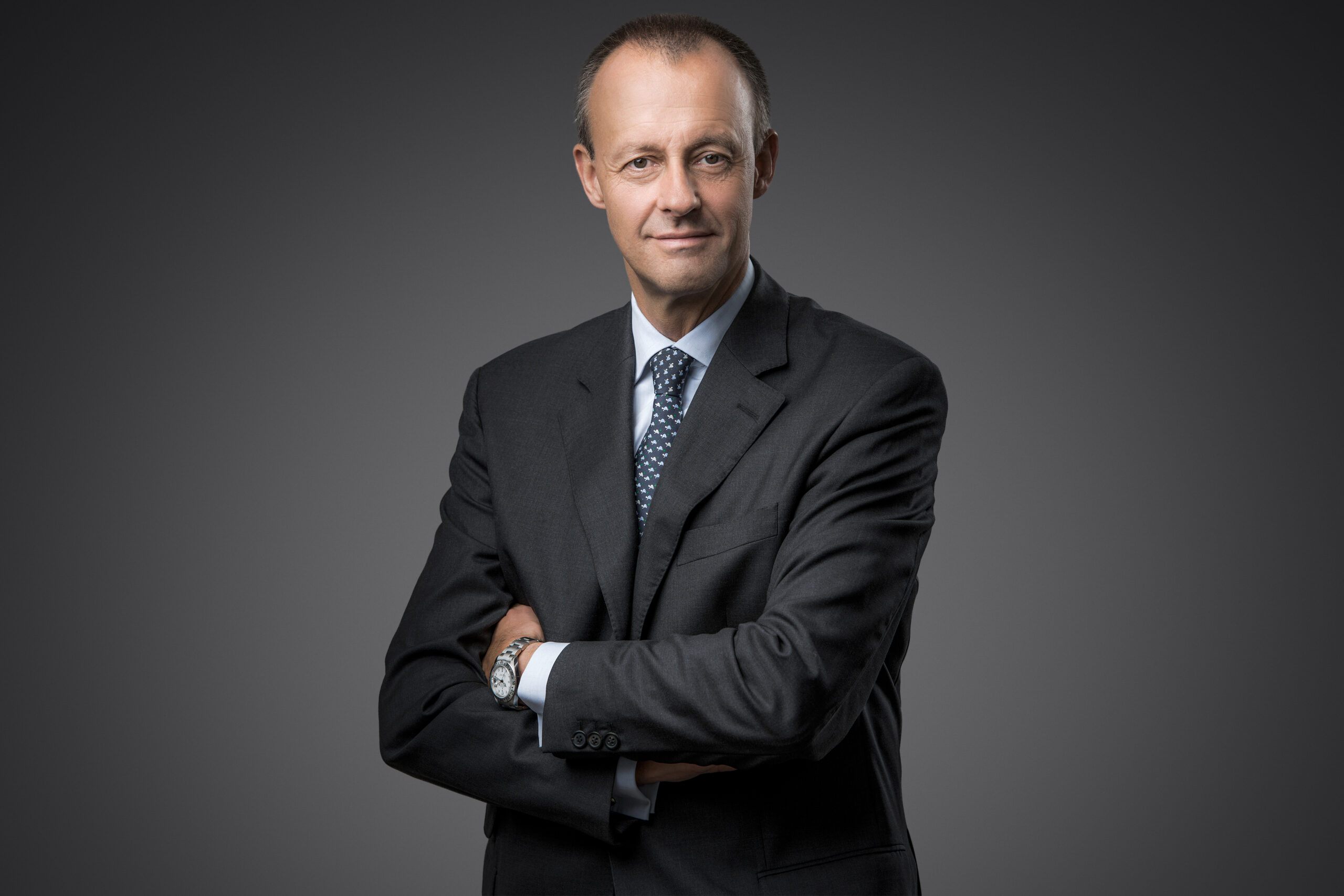 Der Bundeskanzler Friedrich merz steht vor grauem Hintergrund, hat die Arme verschränkt und schaut intensiv in die Kamera.