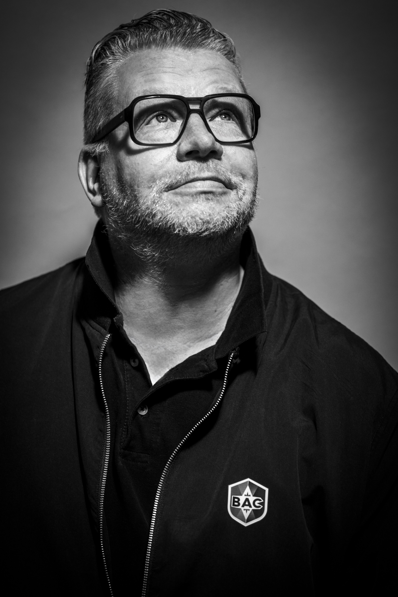 Das Foto des Mannes ist schwarz weiß. Ein Mann schaut nach oben rechts. Er trägt eine schwarze Brille und ein schwarzes Oberteil.