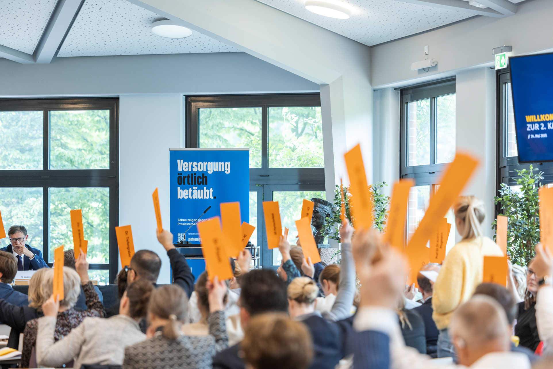 Menschen Stimmen ab in einer Tagung in der Zahnärztekammer Nordrhein. Orangene Zettel werden hochgehalten.